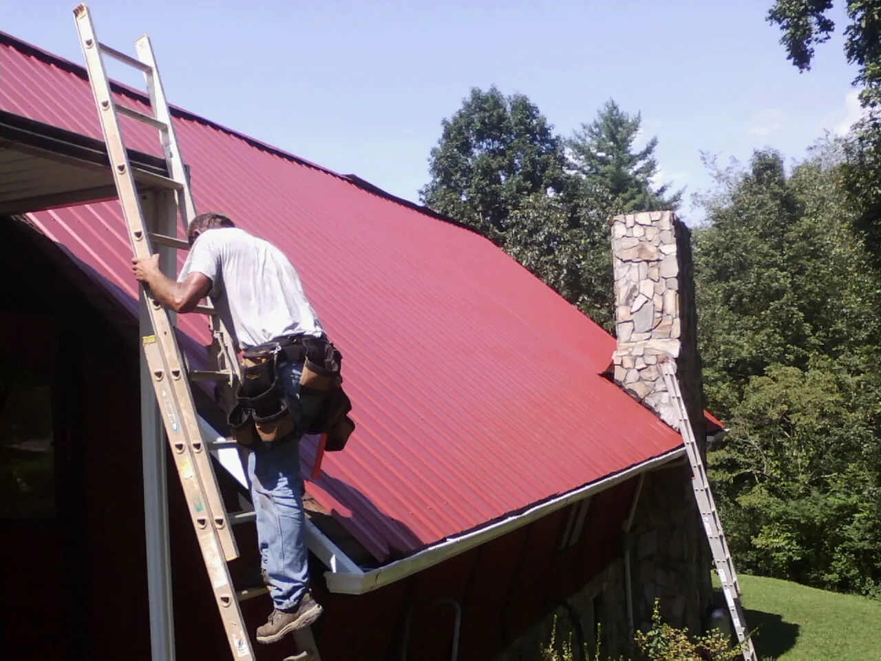 Roofer on a ladder beside a red metal roof and stone chimney — Soffit & Fascia Repair by Roofing Brothers in Walford